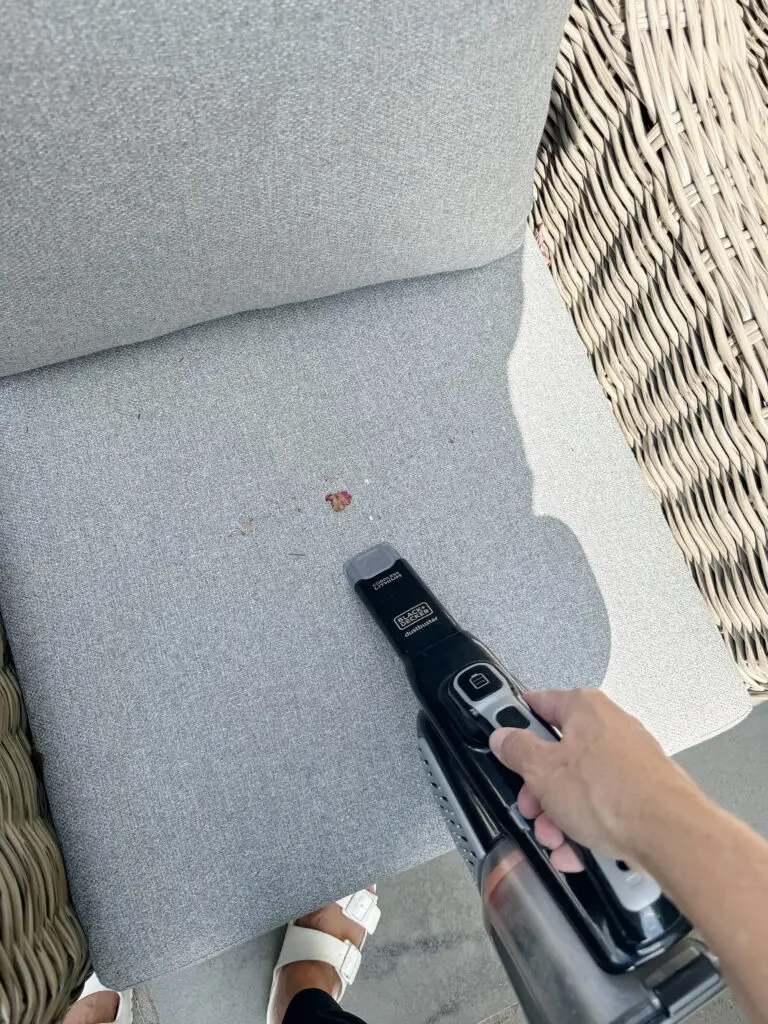 A person uses a handheld vacuum cleaner to remove crumbs from a light gray cushioned chair.