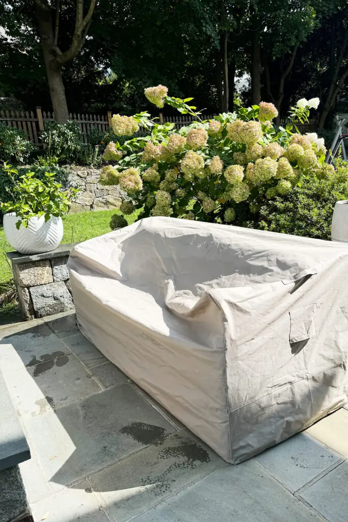 A patio sofa covered with a protective beige tarp sits on a stone patio near blooming hydrangeas and a white planter in a garden setting.