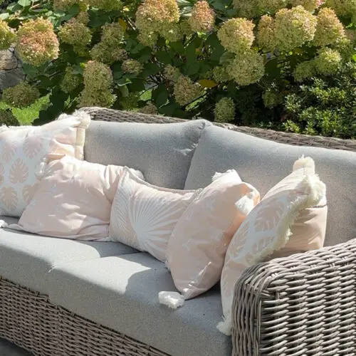 A wicker outdoor sofa with light gray cushions and decorative pillows sits on a stone patio, with hydrangea bushes and greenery in the background.