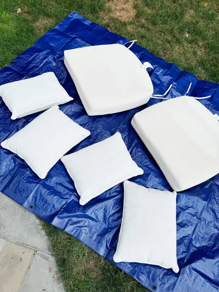 Six white cushions are laid out on a blue tarp on the grass, with some patches of dry grass visible in the background.