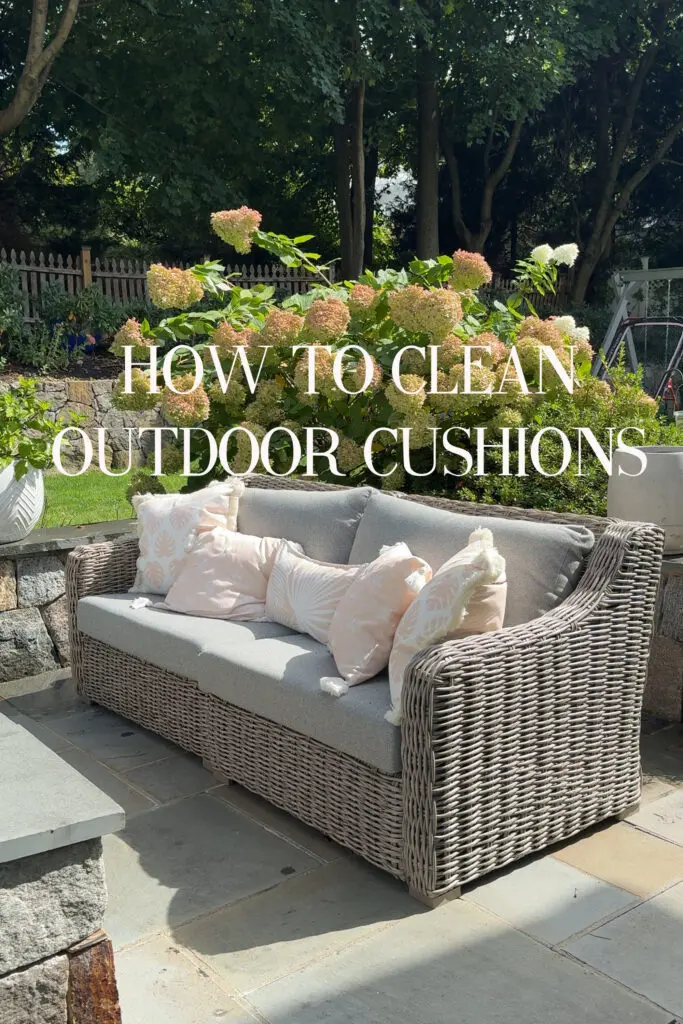 The simple tasks of how to clean outdoor cushions requires just a little elbow grease and a sunny afternoon and you can have your outdoor cushions looking new. A wicker outdoor sofa with light gray cushions and pastel pillows sits on a stone patio in a sunny garden. Text reads: "How to clean outdoor cushions.