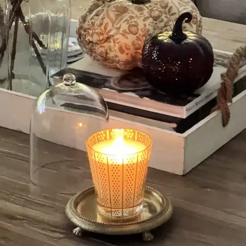 A lit candle in a decorative holder sits on a small tray with a glass cloche beside it, next to a tray holding two decorative pumpkins and two glass bottles on a wooden table.