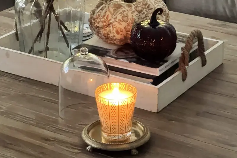 A lit candle in a decorative holder sits on a small tray with a glass cloche beside it, next to a tray holding two decorative pumpkins and two glass bottles on a wooden table.
