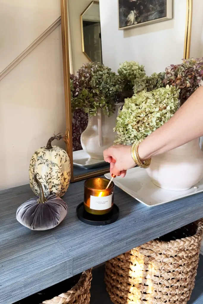 A person lights a candle on a table decorated with artificial pumpkins, vases of dried hydrangeas, and a large mirror.