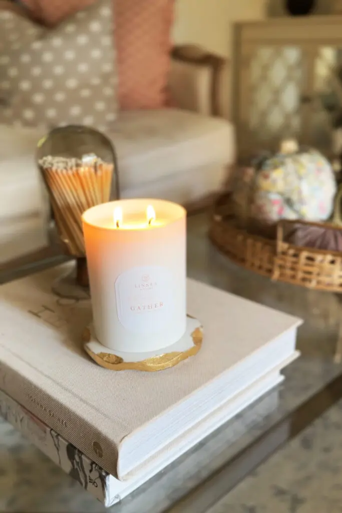 A lit white candle labeled "Gather" sits on top of stacked books on a glass coffee table, with a bowl of matches and a decorative tray in the background.