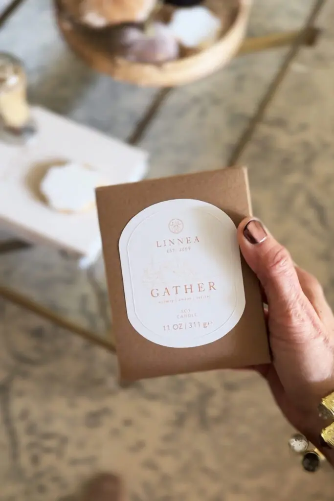 A hand holds a brown box labeled "Linnea Gather Candle, 11 oz / 311 g," with a blurred table and candle in the background.