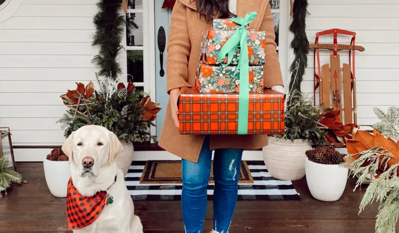 A person in a brown coat holds stacked wrapped gifts, ready to consult the 2025 Holiday Gift Guides, while standing next to a yellow lab in a plaid bandana on a festively decorated porch.