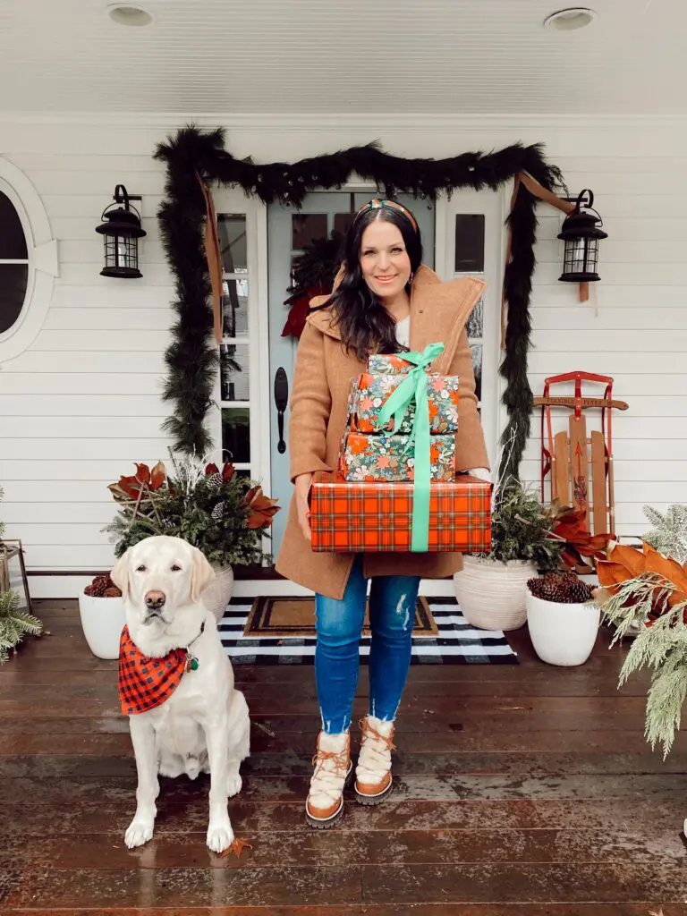 A woman in a brown coat holds a stack of wrapped gifts on a porch decorated for the holidays. A dog wearing a red scarf sits beside her.