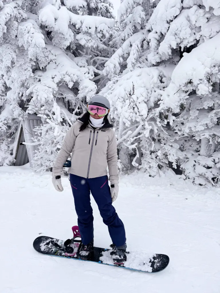 Person wearing winter gear stands on a snowboard in a snowy landscape with snow-covered trees in the background.