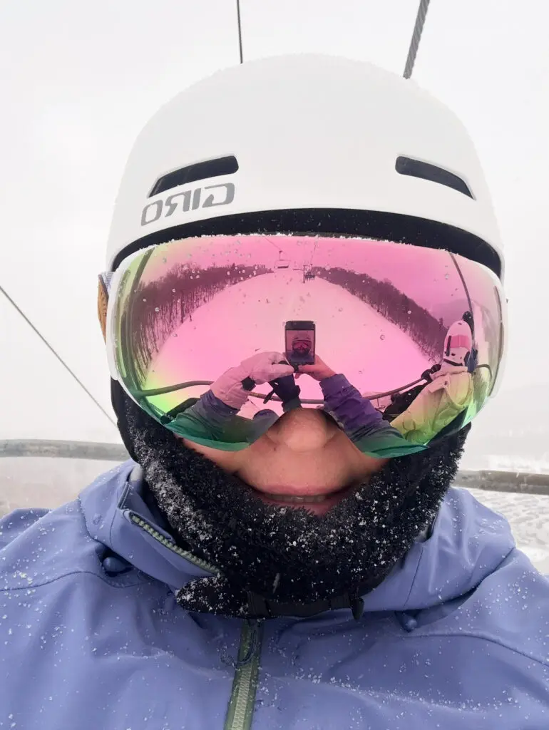 Person wearing a white helmet and reflective ski goggles takes a selfie on a ski lift in snowy weather. Their reflection and phone are visible in the goggles.