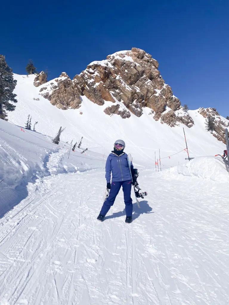 A person in ski gear stands on a snowy slope, holding skis with a rocky, snow-covered mountain and clear blue sky in the background.