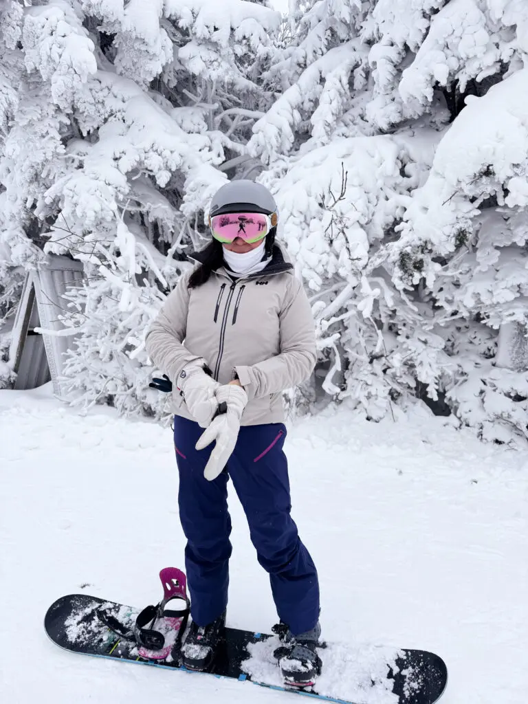 A person in winter sports gear stands on a snowboard in a snowy, tree-filled landscape.