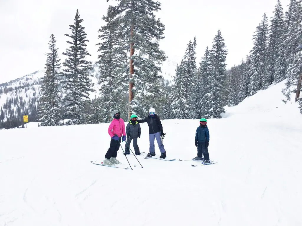 Four people wearing winter gear stand on a snowy ski slope surrounded by tall pine trees and mountains in the background.