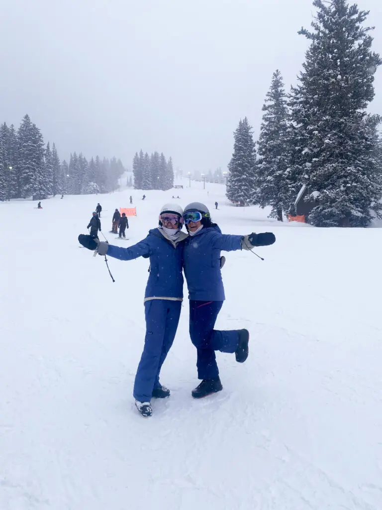 Two people in ski gear pose together on a snowy ski slope with trees and other skiers in the background on a cloudy day.