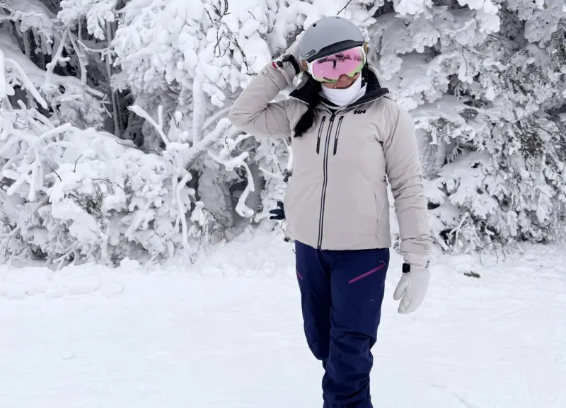 A person in winter clothing and a helmet stands in the snow near snow-covered trees, adjusting their ski goggles.