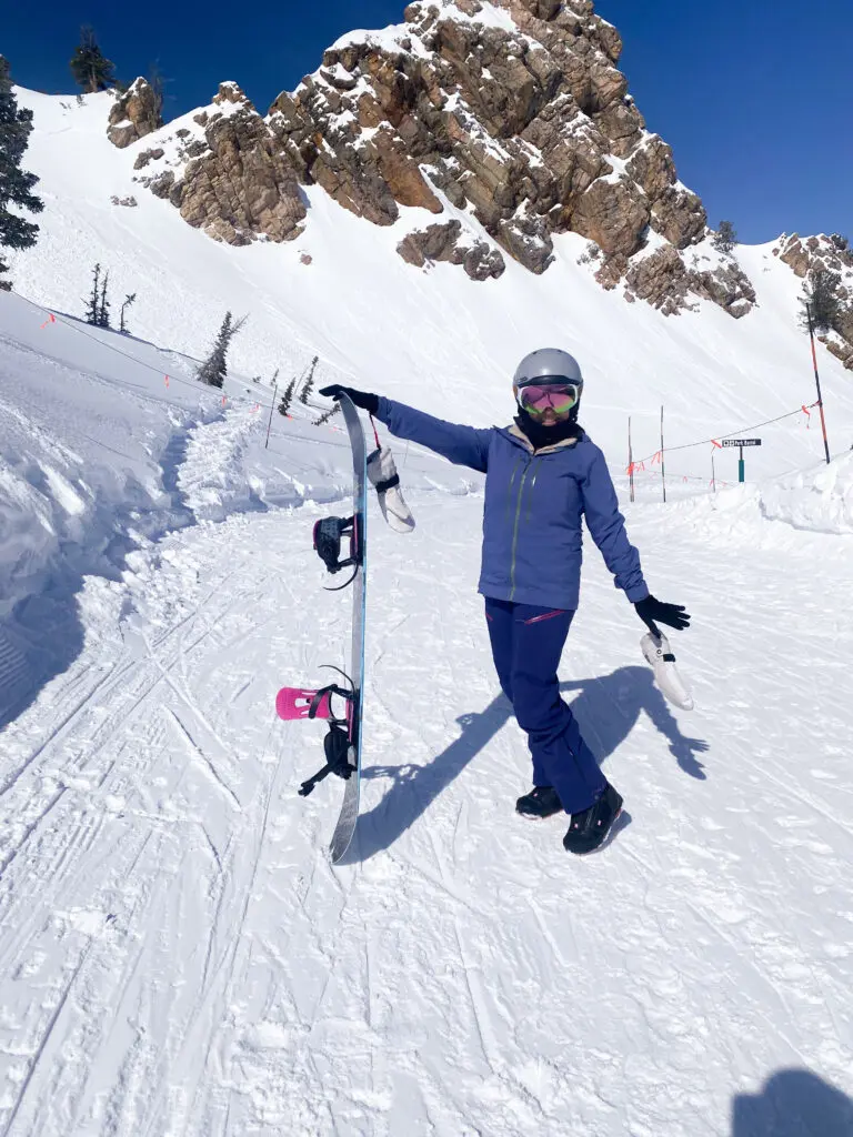 Person in winter gear poses on a snowy mountain slope, holding a snowboard vertically. Rocky cliffs and ski trail markers are visible in the background under a clear blue sky.