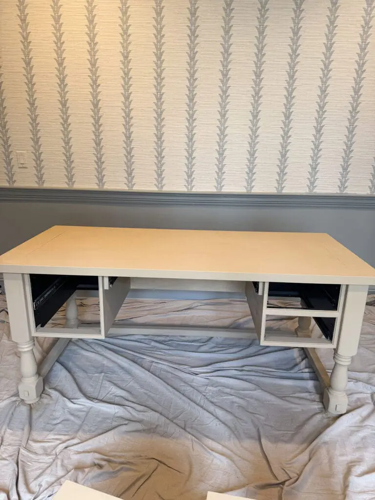 DIY Pottery Barn Finish Furniture || Darling Darleen Top Lifestyle Blogger A partially assembled white desk sits on a drop cloth in a room with patterned wallpaper and gray wainscoting.