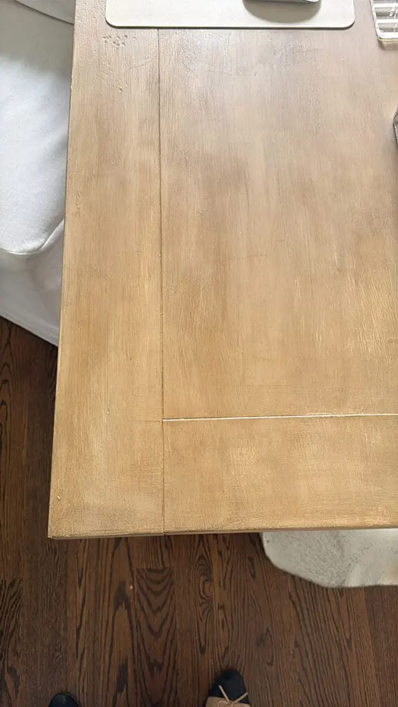 A wooden desk with a visible corner seam, placed on a wooden floor near a white upholstered chair. Part of a person's feet and a mouse pad are also visible.