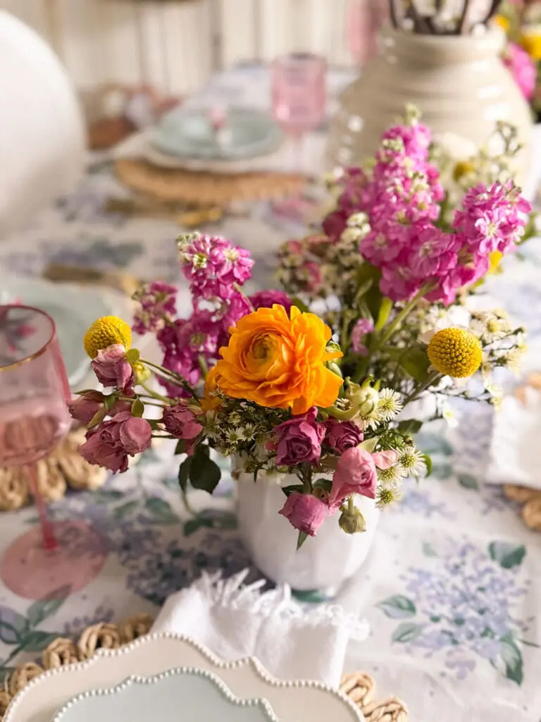 A white vase with a bouquet of yellow, pink, and purple flowers sits on a floral tablecloth, surrounded by pastel plates and pink glassware.