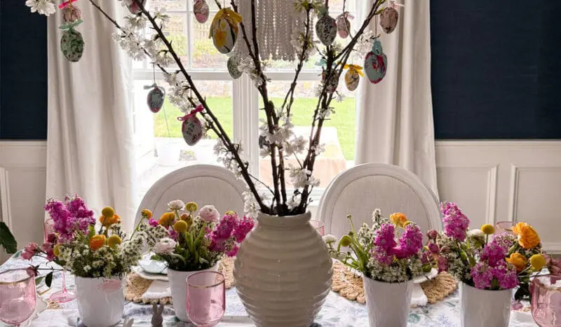 A dining table is decorated with spring flowers in vases and a large white vase with blossoming branches adorned with hanging Easter egg ornaments.