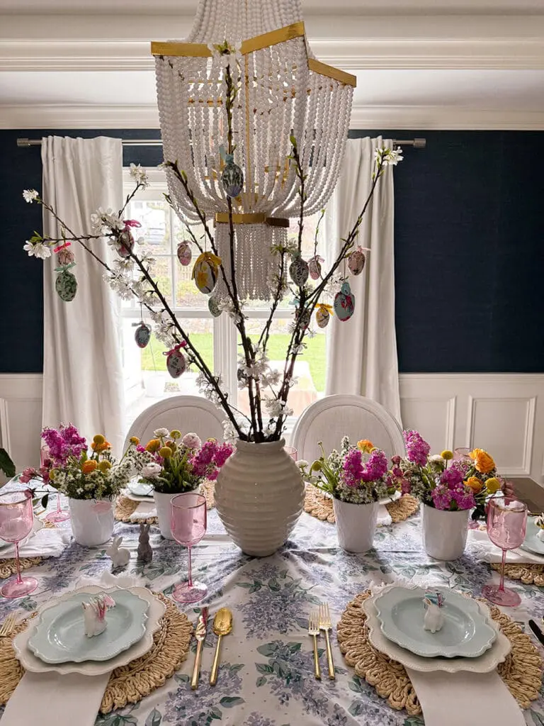 A dining table set for a meal, decorated with a floral tablecloth, pink glasses, flower centerpieces, and a large vase with branches and hanging ornaments under a beaded chandelier.