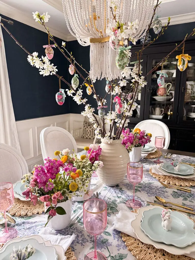 A dining table set for spring with floral arrangements, pink glassware, and a centerpiece of branches decorated with hanging Easter eggs.