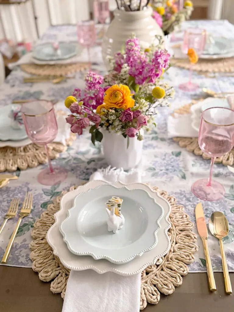A dining table set with pastel plates, pink glasses, gold cutlery, woven placemats, and floral centerpieces in white vases. A white napkin with a bunny-shaped holder is on each plate.