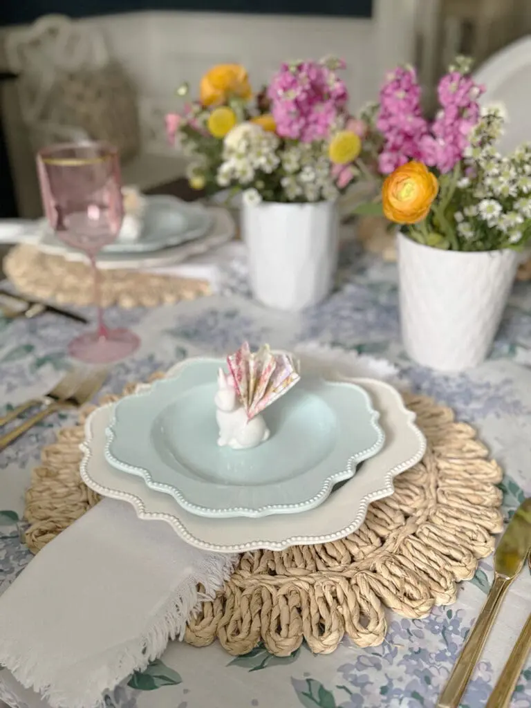 A table is set with layered white and blue plates, a white napkin, a pink goblet, and a small bunny figurine holding a folded paper on a woven placemat, with flowers in vases.