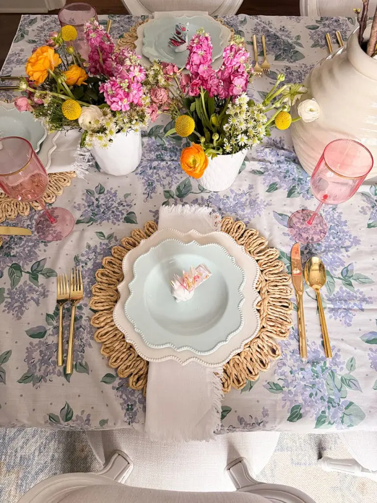A spring-themed table setting with floral placemats, gold cutlery, scalloped plates, pink glasses, and flower arrangements in white vases.