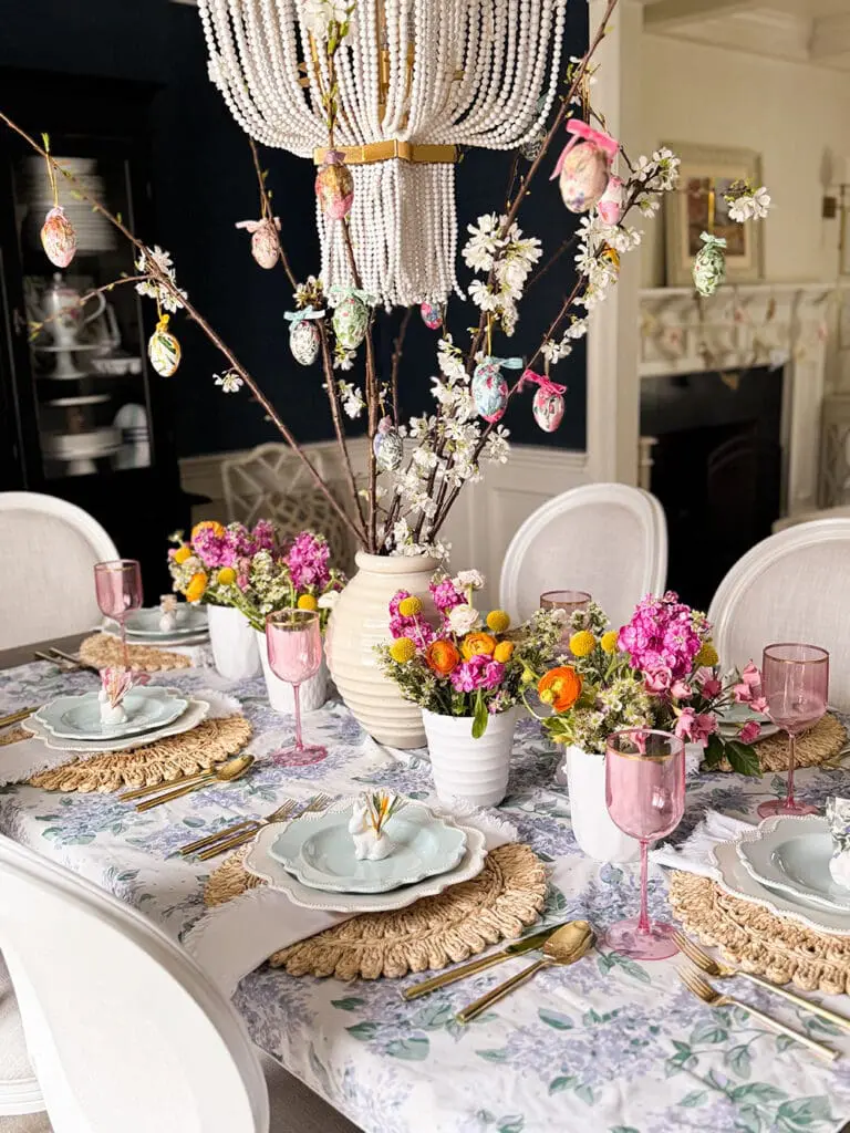 Elegant dining table set for spring with floral arrangements, pink glassware, pastel plates, and a centerpiece of branches decorated with colorful hanging eggs beneath a beaded chandelier.