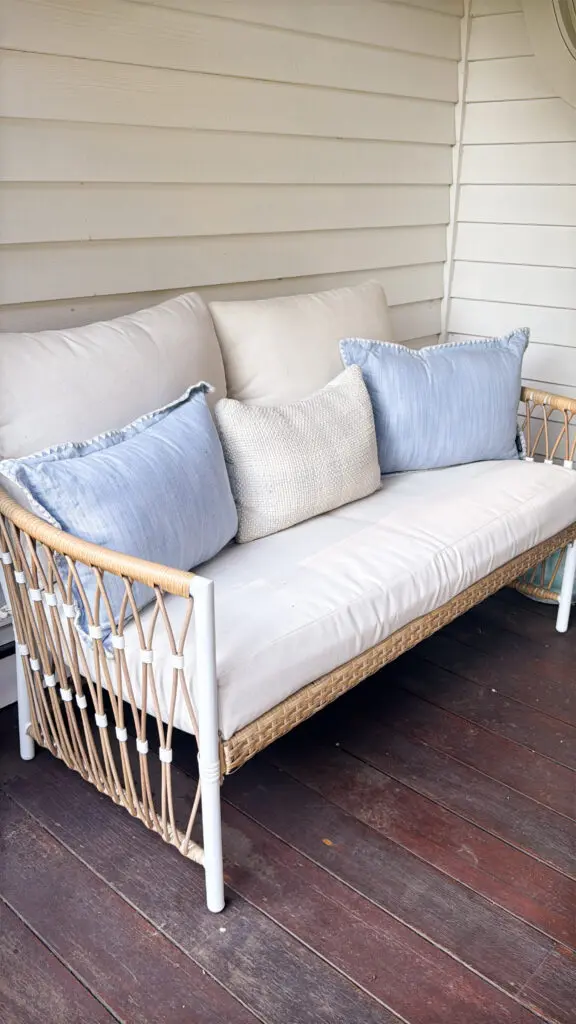 A wicker and metal framed outdoor sofa with white cushions and three pillows, two blue and one beige, placed on a wooden porch against a white paneled wall.