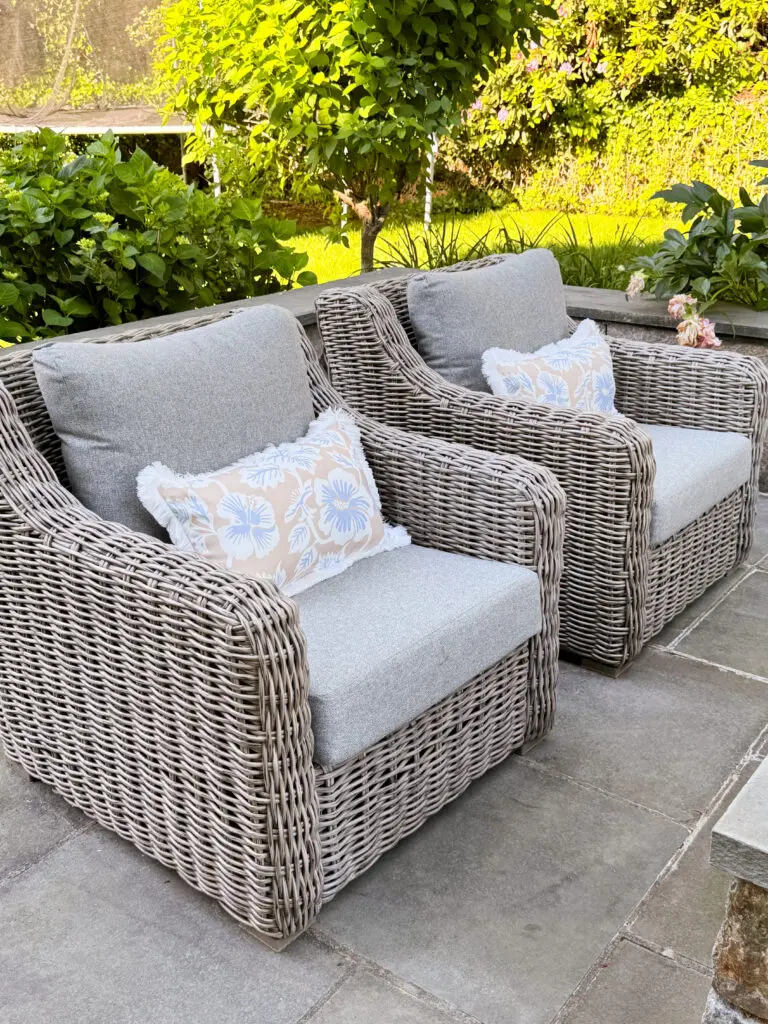 Two wicker patio chairs with gray cushions and floral-patterned pillows are placed side by side on a stone patio surrounded by green plants.