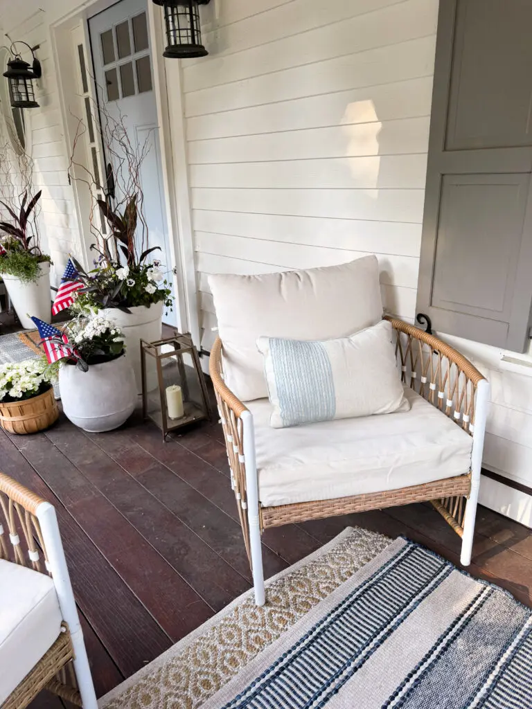 Wicker chair with cream cushions and a striped pillow on a porch with potted plants, small American flags, and a striped rug, next to a white house wall and door.