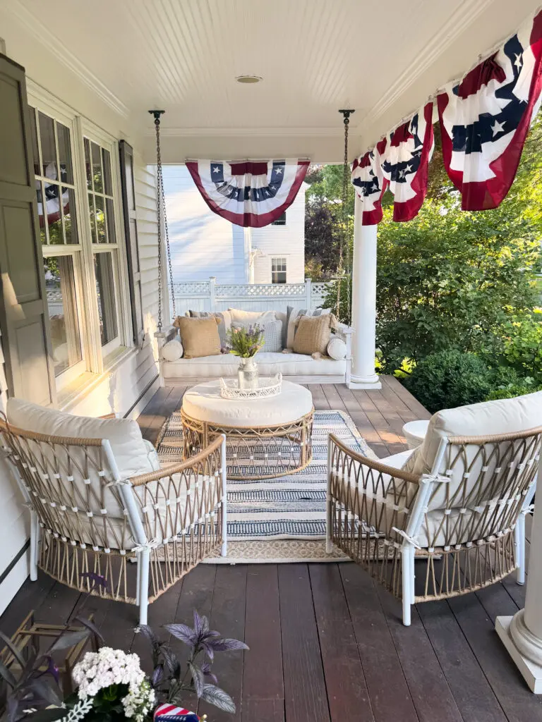 A cozy front porch with wicker furniture, a striped rug, a porch swing, and patriotic red, white, and blue bunting hanging from the ceiling.
