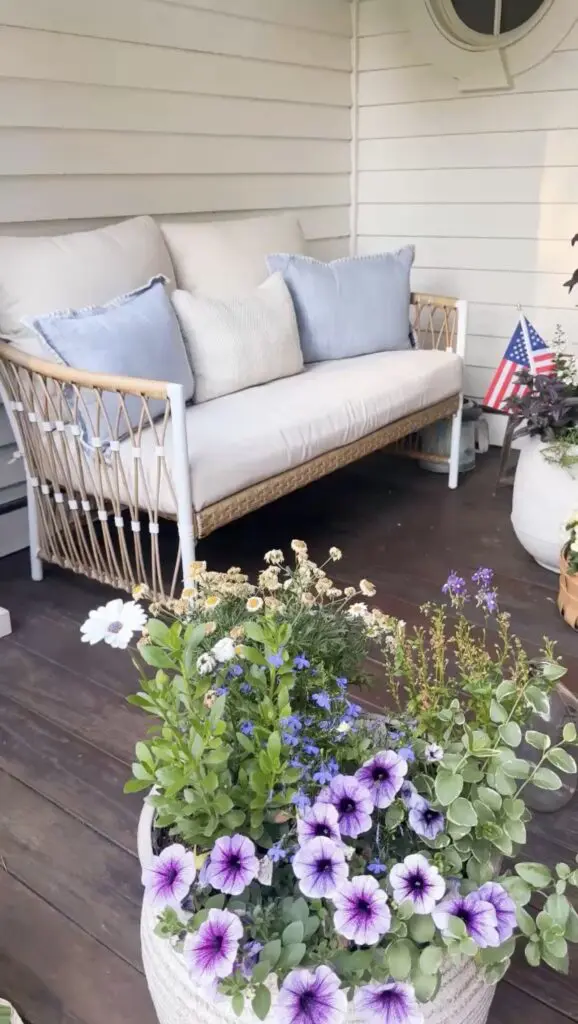 A white outdoor sofa with blue cushions sits on a wooden deck, surrounded by potted flowers and an American flag.