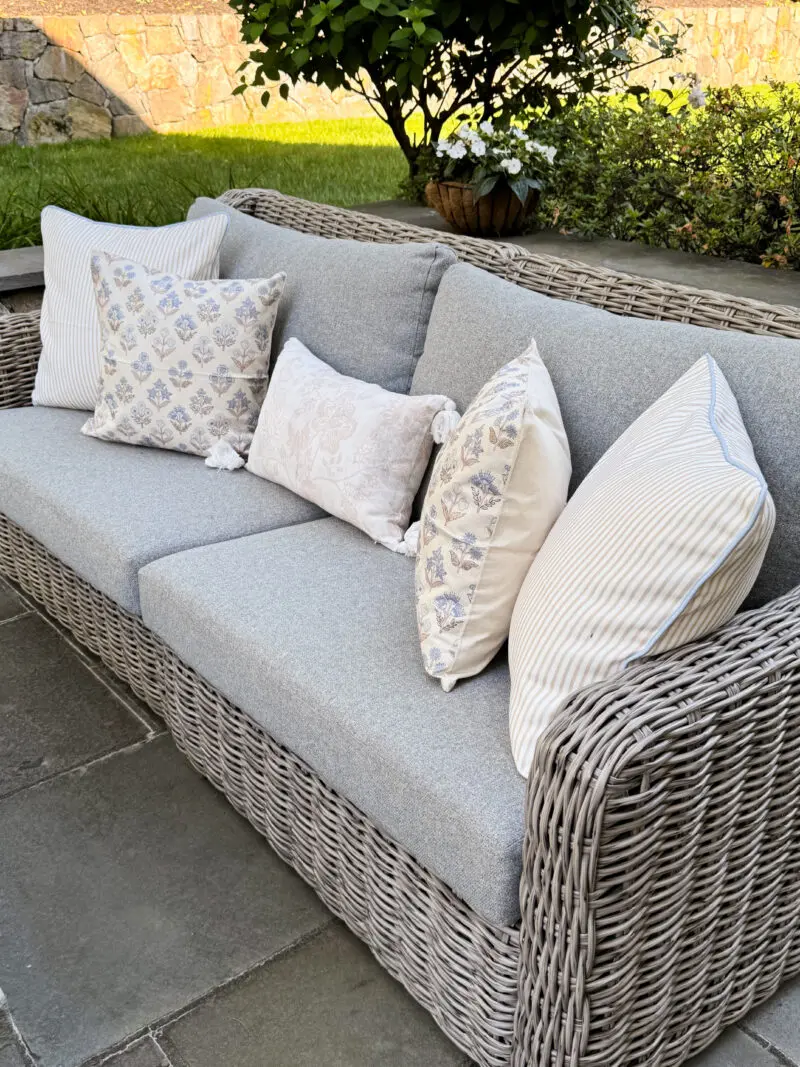 Grey wicker outdoor sofa with light grey cushions and five patterned decorative pillows, placed on a stone patio with greenery and a stone wall in the background.