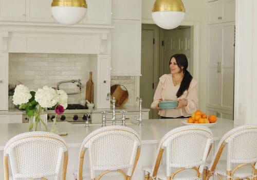 A woman in a beige blouse smiles while holding a blue bowl in a bright kitchen with white cabinets and gold pendant lights.