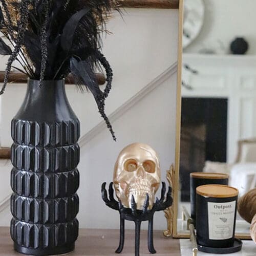 A halloween-themed mantel decor featuring a golden skull, black feathered branches in a vase, pumpkins, and a candle.