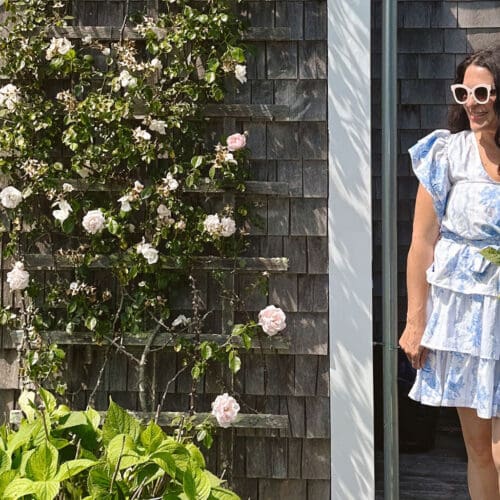 A woman in a blue and white dress smiling and holding a large bouquet of blue flowers next to a gray wooden house with climbing roses.