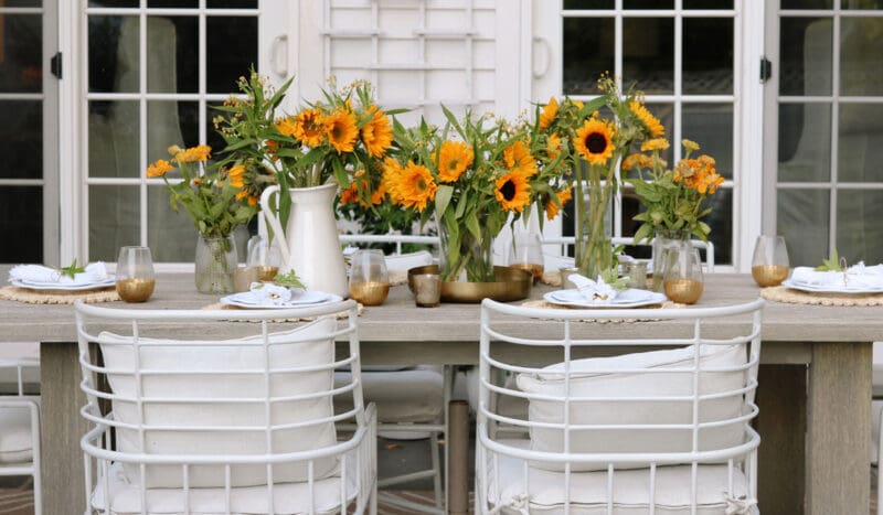 Outdoor dining setup with white chairs and wooden tables decorated with sunflowers in vases and white dinnerware.