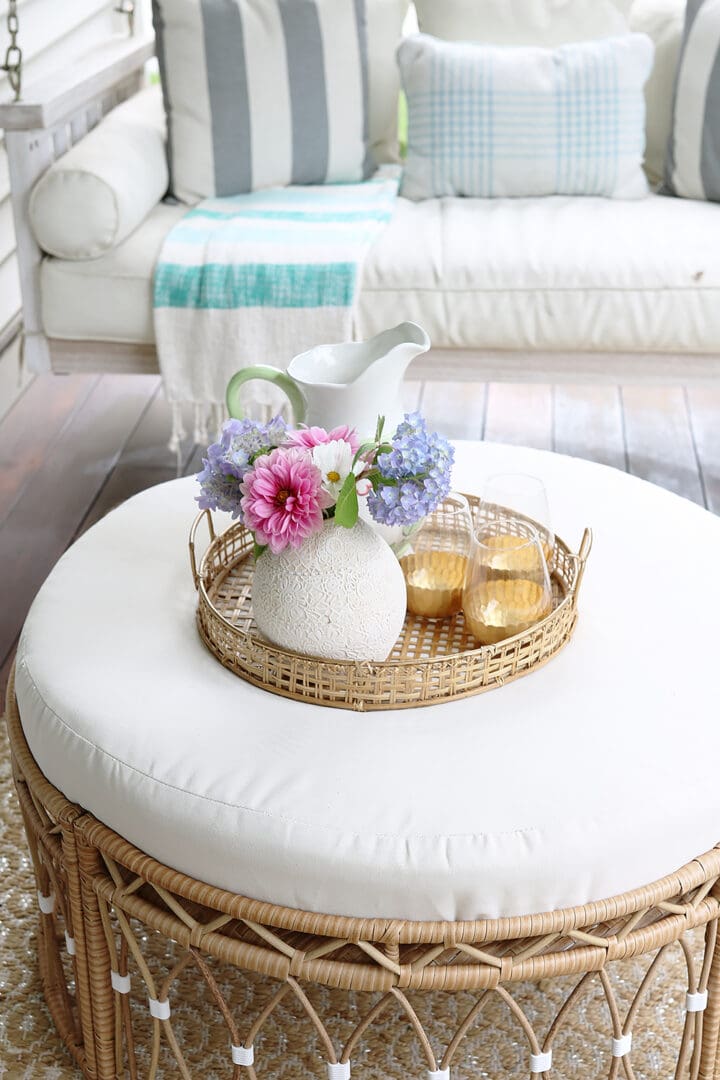 Rattan ottoman with a wicker tray holding a vase of flowers, two glasses of juice, and a pitcher, set on a patio with a white cushioned seating area in the background.