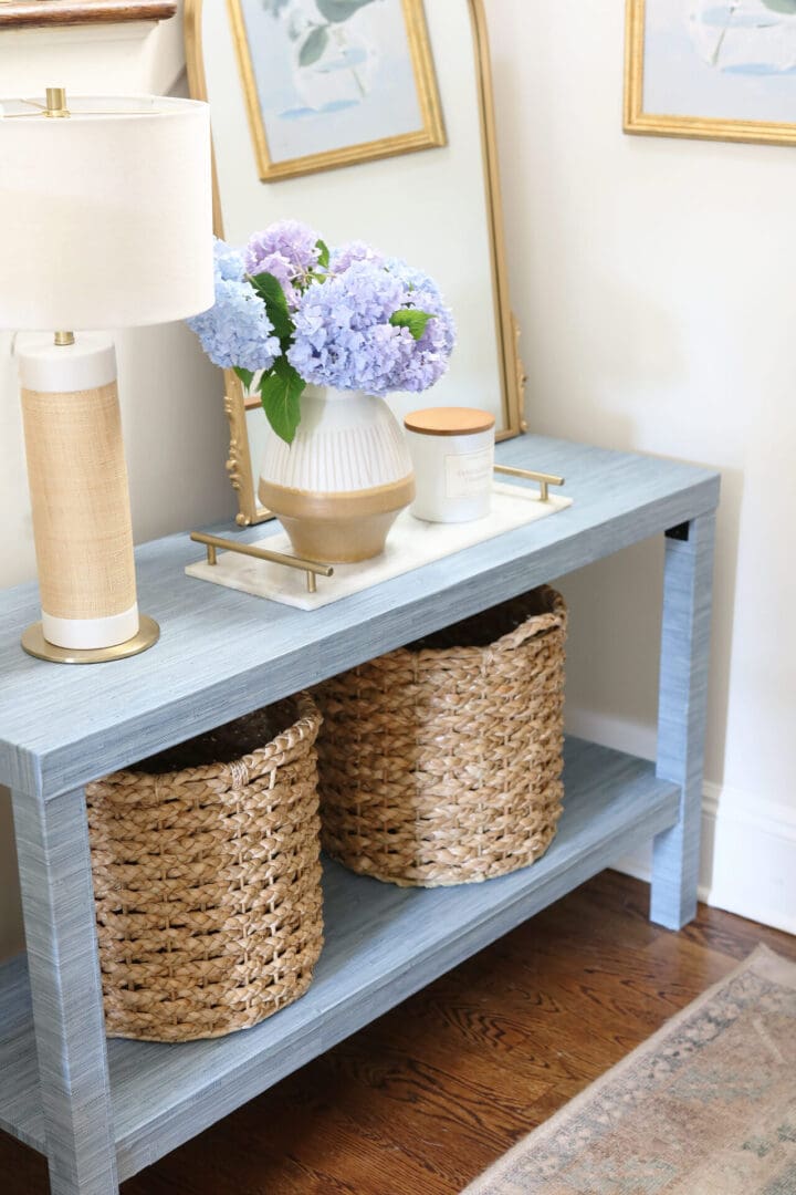 A blue wooden console table with a lamp, a vase of purple flowers, a candle, and a mirror on top. Below the table are two woven baskets. The table is placed against a light-colored wall.