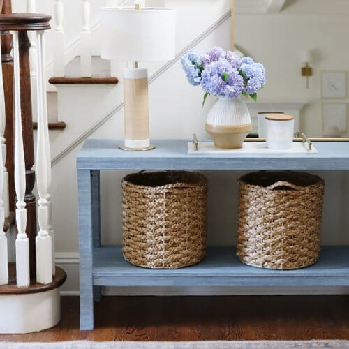 A hallway features a blue console table with two woven baskets underneath, a table lamp, a vase with hydrangeas, and a candle. The table is set against a staircase with white railings and wooden steps.