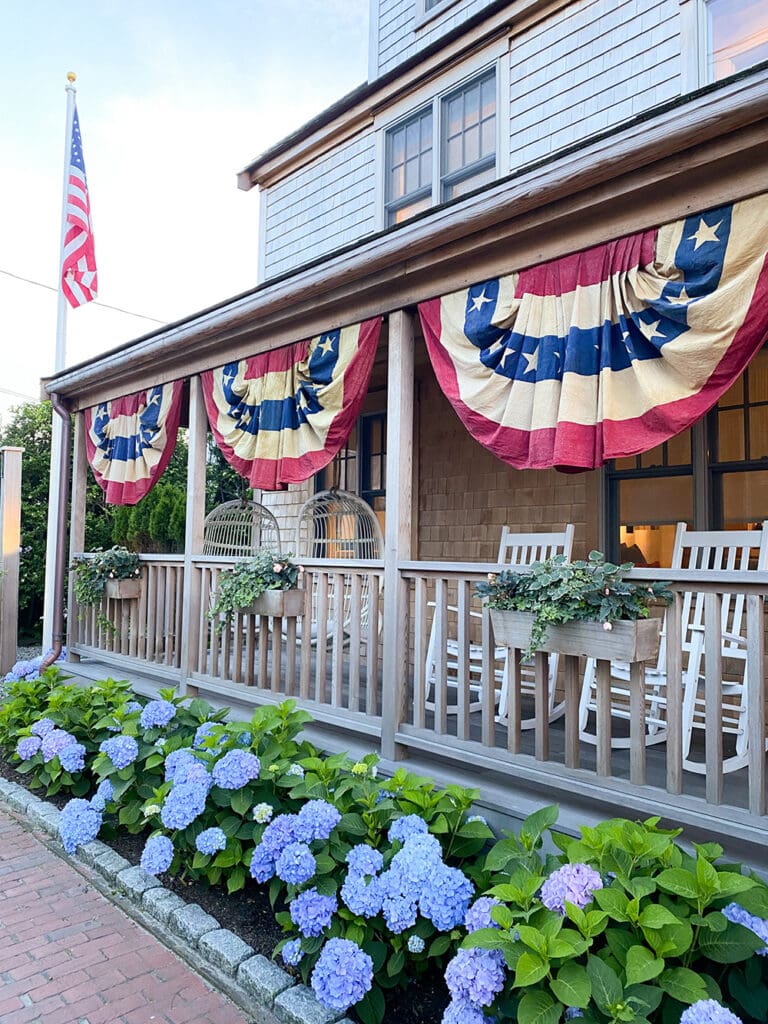 Fourth of July in Nantucket: A Nantucket porch decorated with American flag bunting and rocking chairs. Hydrangea flowers bloom along the brick pathway in front of the porch. An American flag is displayed on a pole.