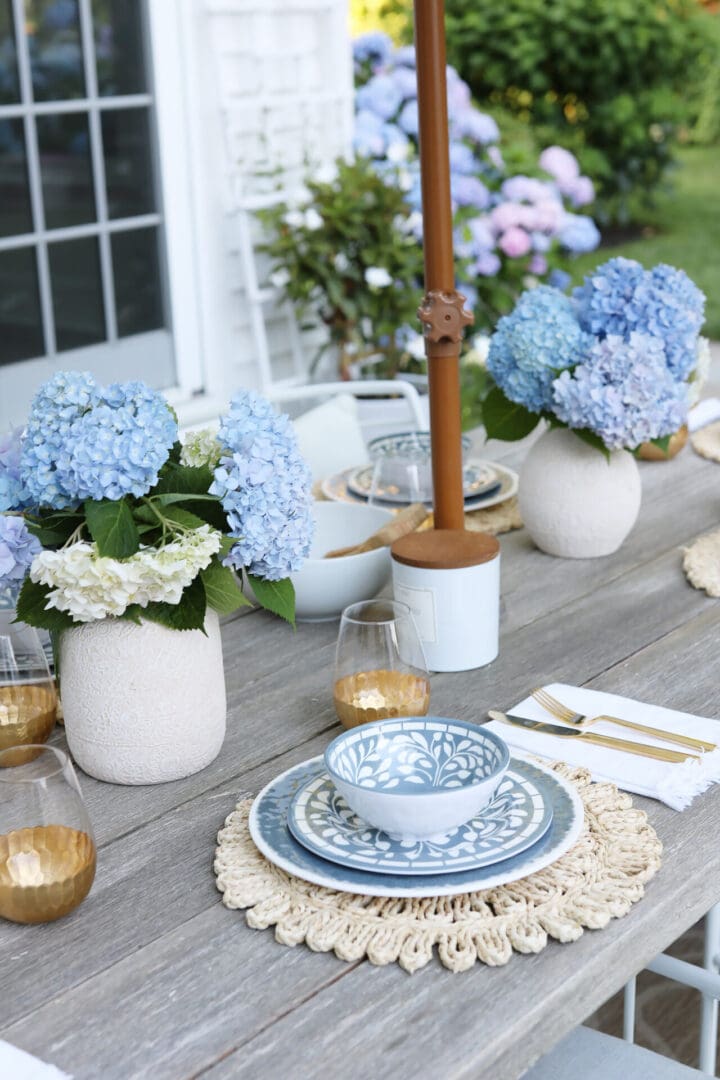 A wooden table is set for a meal with blue and white patterned dishes, cutlery, gold-rimmed glasses, and vases of blue hydrangeas.