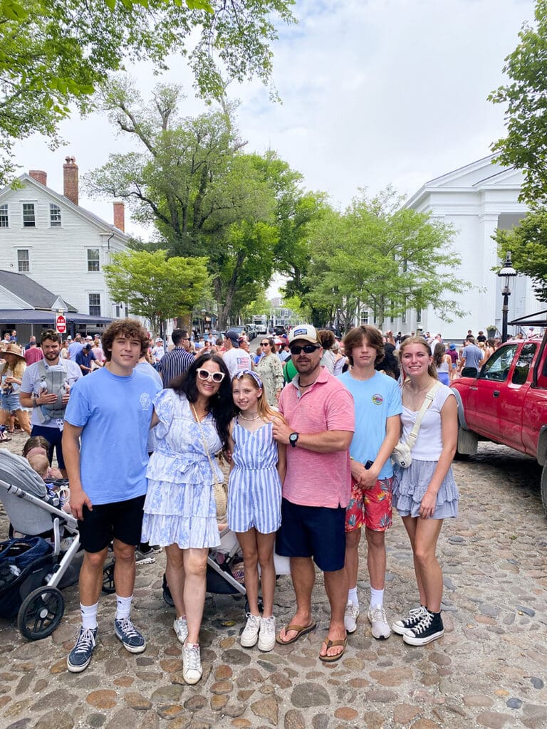 : Fourth of July in NantucketA family of six poses on a cobblestone street during a busy outdoor event. Several people and trees are visible in the background.