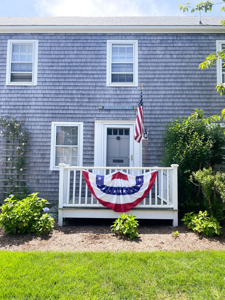 Fourth of July in Nantucket: A gray shingled house with a white door and two windows. An American flag and a patriotic red, white, and blue decoration adorn the porch railing. Green plants and a lawn are in front.