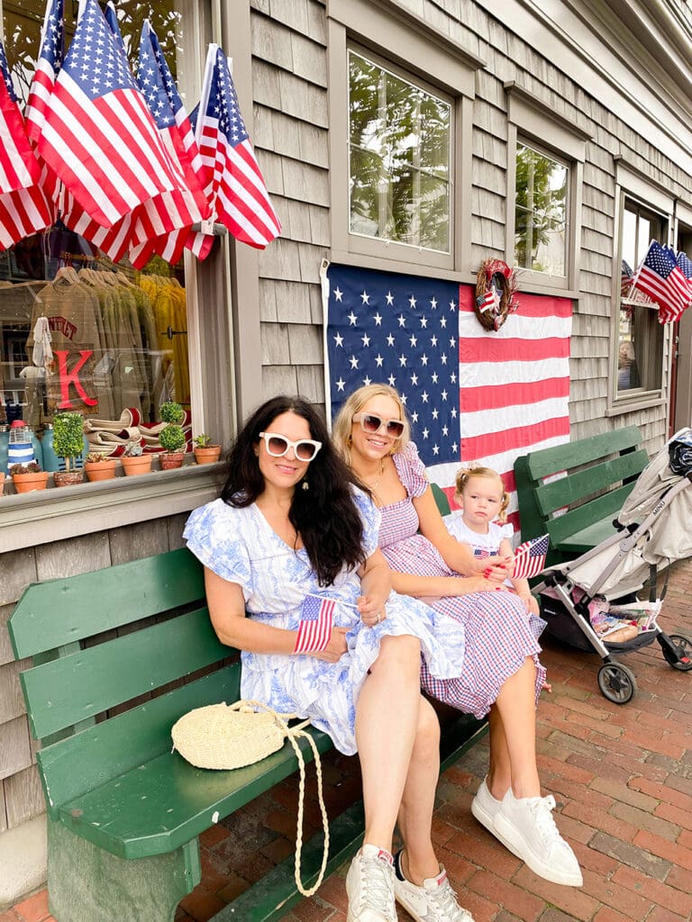 Fourth of July in Nantucket: Three people, two adults and one child, sit on a bench. They are dressed in red, white, and blue clothing and hold small American flags. Behind them, there are larger American flags and a flag-themed decor on a building.