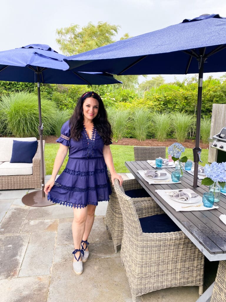 Fourth of July in Nantucket: Woman in a navy dress stands by an outdoor dining table with a blue umbrella, set with plates and glasses. She is on a patio with greenery in the background.