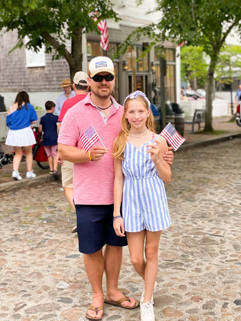 Fourth of July in Nantucket: A man and a girl stand outside on a cobblestone path, both holding small American flags. They are part of a larger group attending an outdoor event. Trees and buildings are in the background.